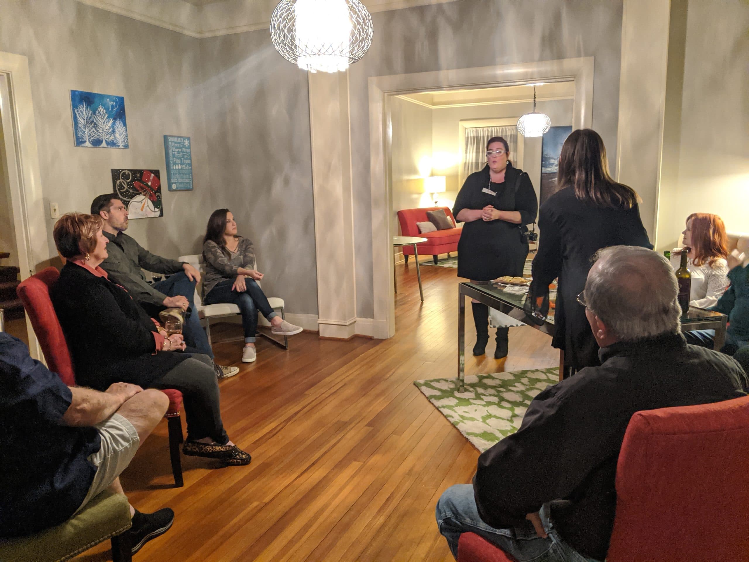 A group of people sits in a cozy living room while a woman stands and speaks at the front.