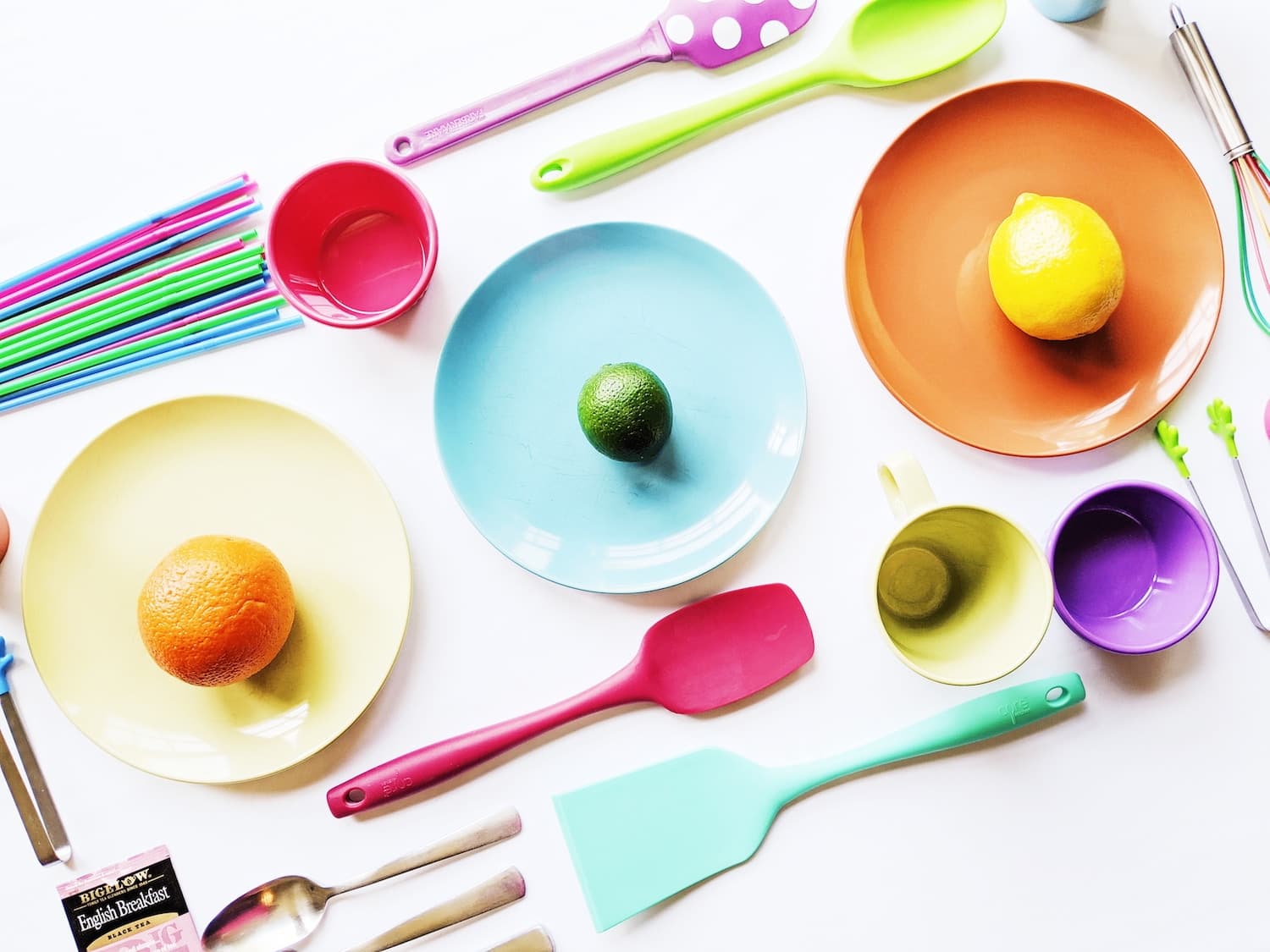 Colorful kitchenware including plates, spoons, and fruit arranged on a white surface.
