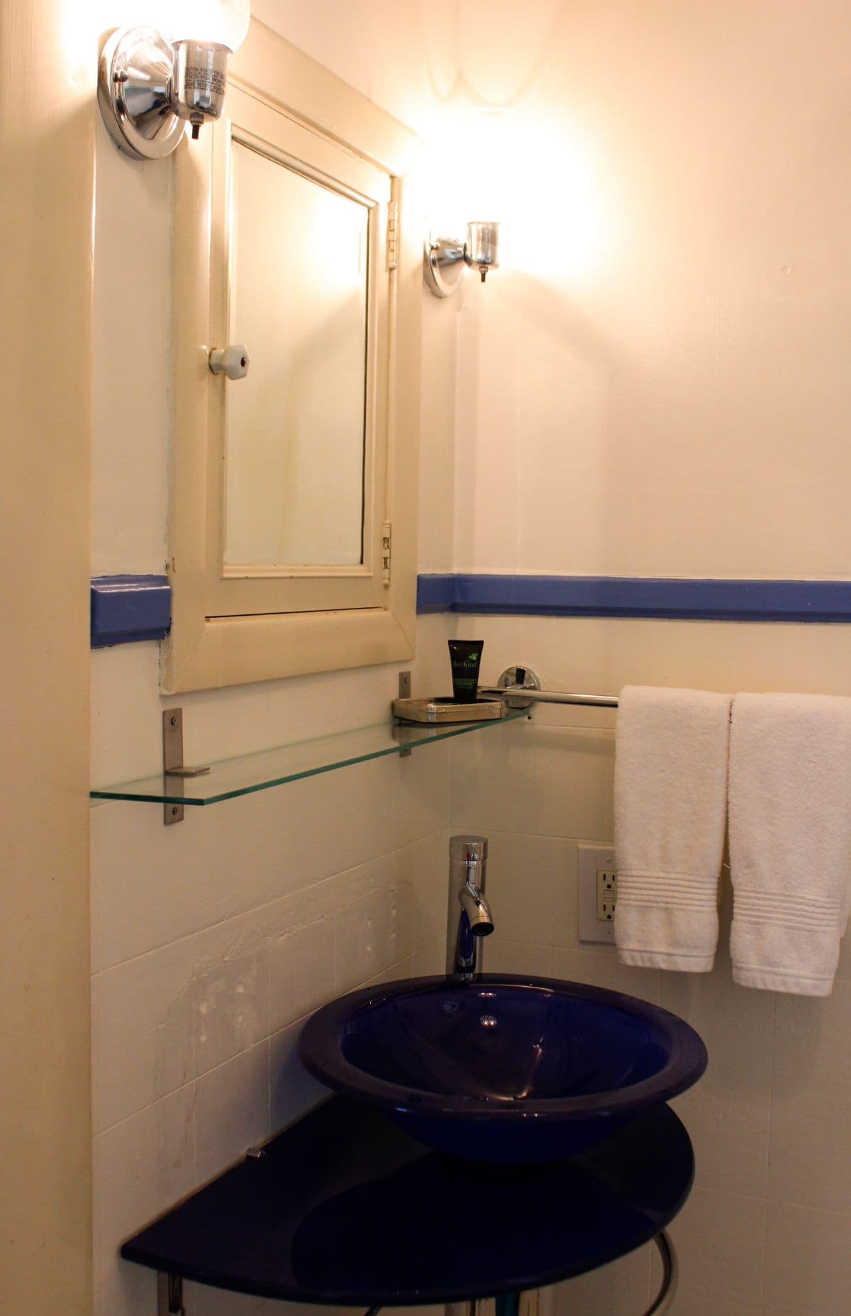 A small bathroom featuring a blue sink, a mirror, a shelf with toiletries, and white towels.