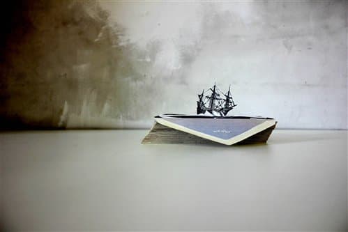 A paper boat with a small ship sculpture sits on a flat surface against a textured wall.