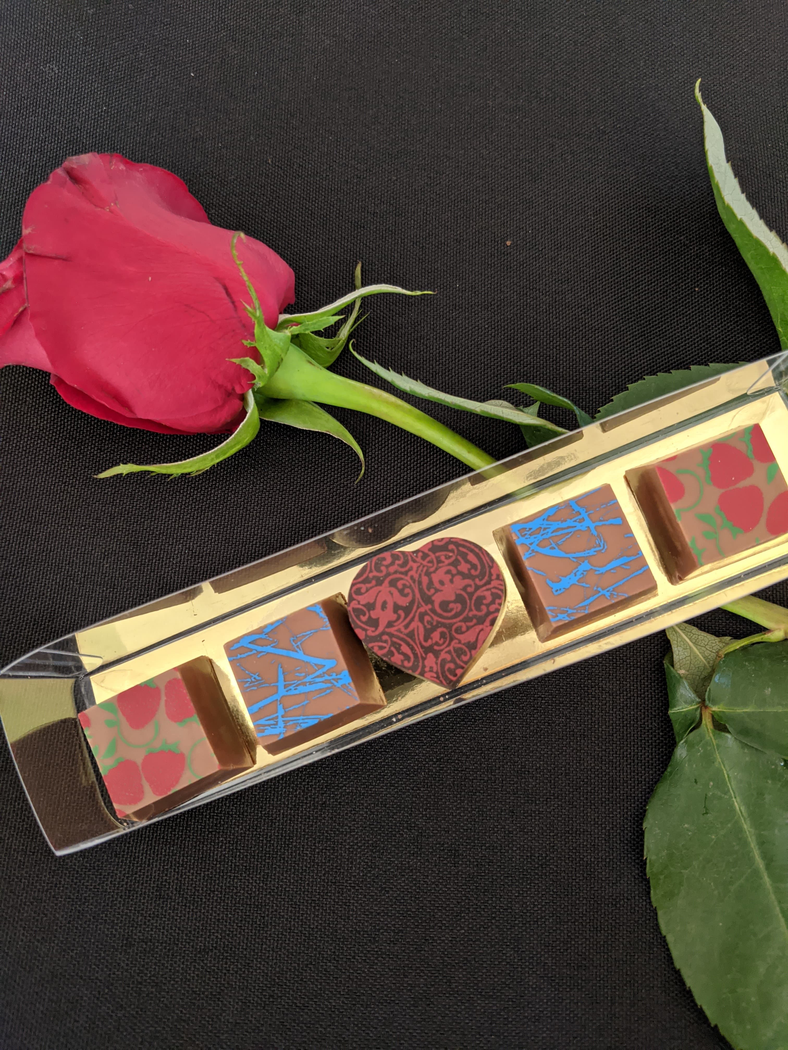 A red rose next to a box of decorative chocolates featuring various designs, resting on a black surface.