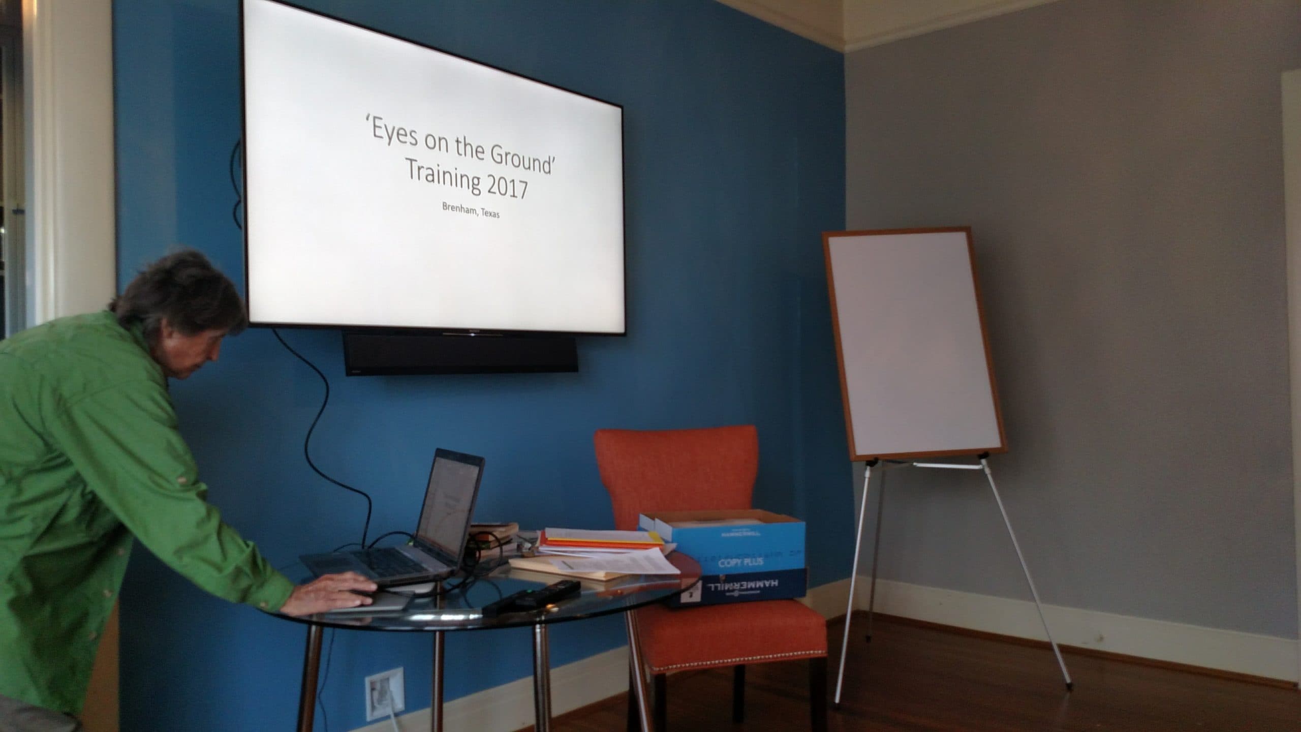A person adjusts a laptop next to a screen displaying "Eyes on the Ground Training 2017" in a conference room.