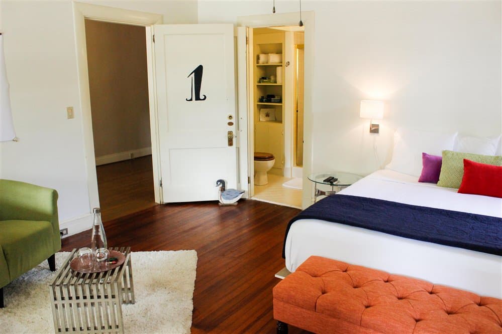 A modern hotel room featuring a bed with colorful pillows, an orange ottoman, and an ensuite bathroom visible through a doorway.