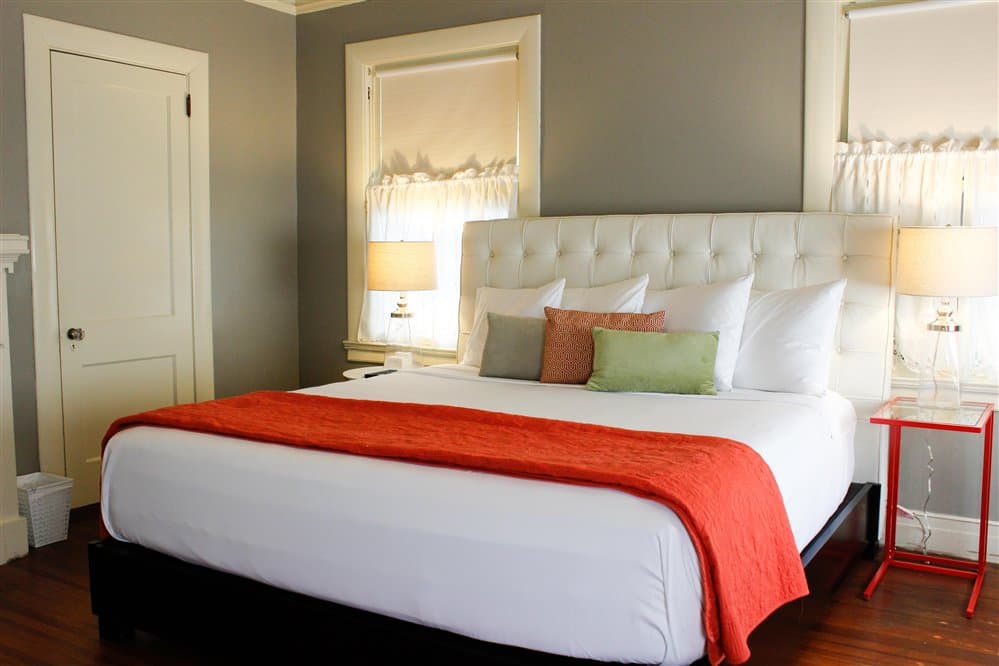 A cozy bedroom featuring a white tufted bed with decorative pillows, orange throw, and warm lighting from bedside lamps.