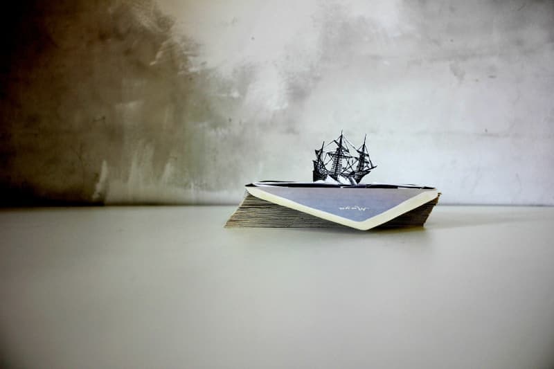 A miniature ship is perched atop a stack of books on a plain surface.