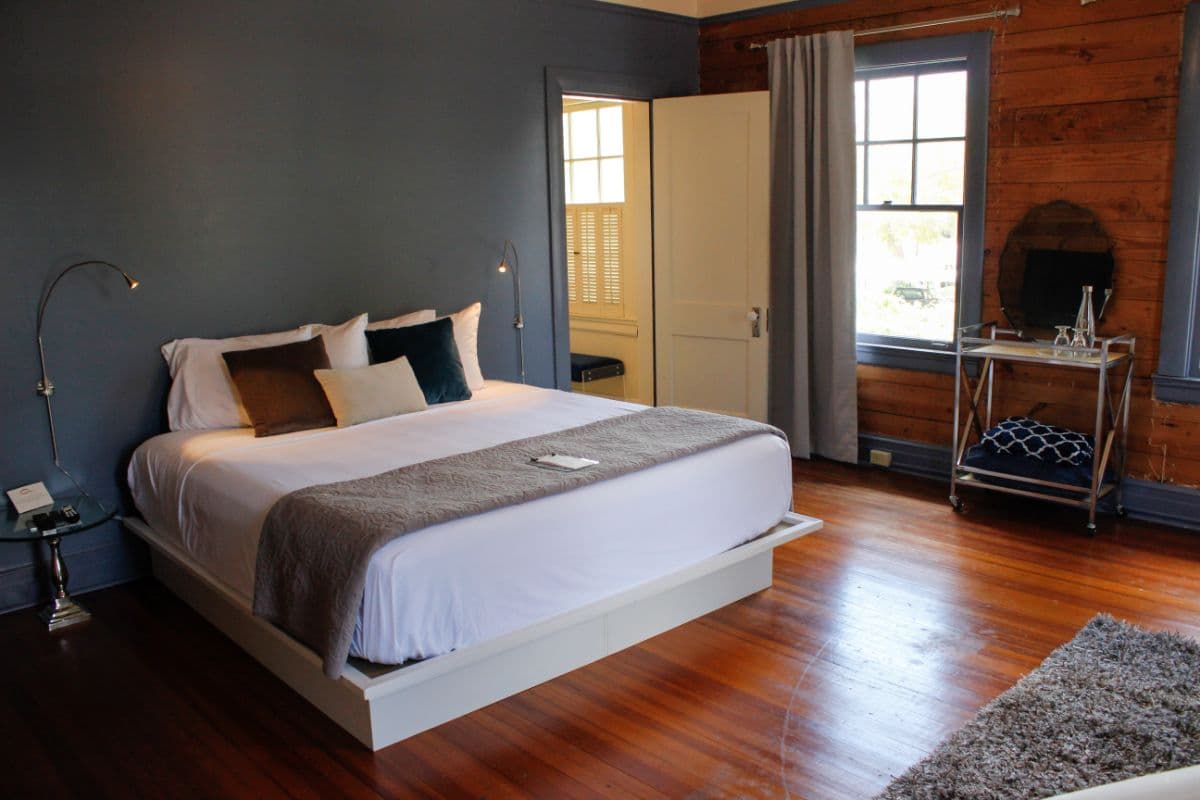 A cozy bedroom featuring a modern bed with decorative pillows, wooden floors, and large windows.