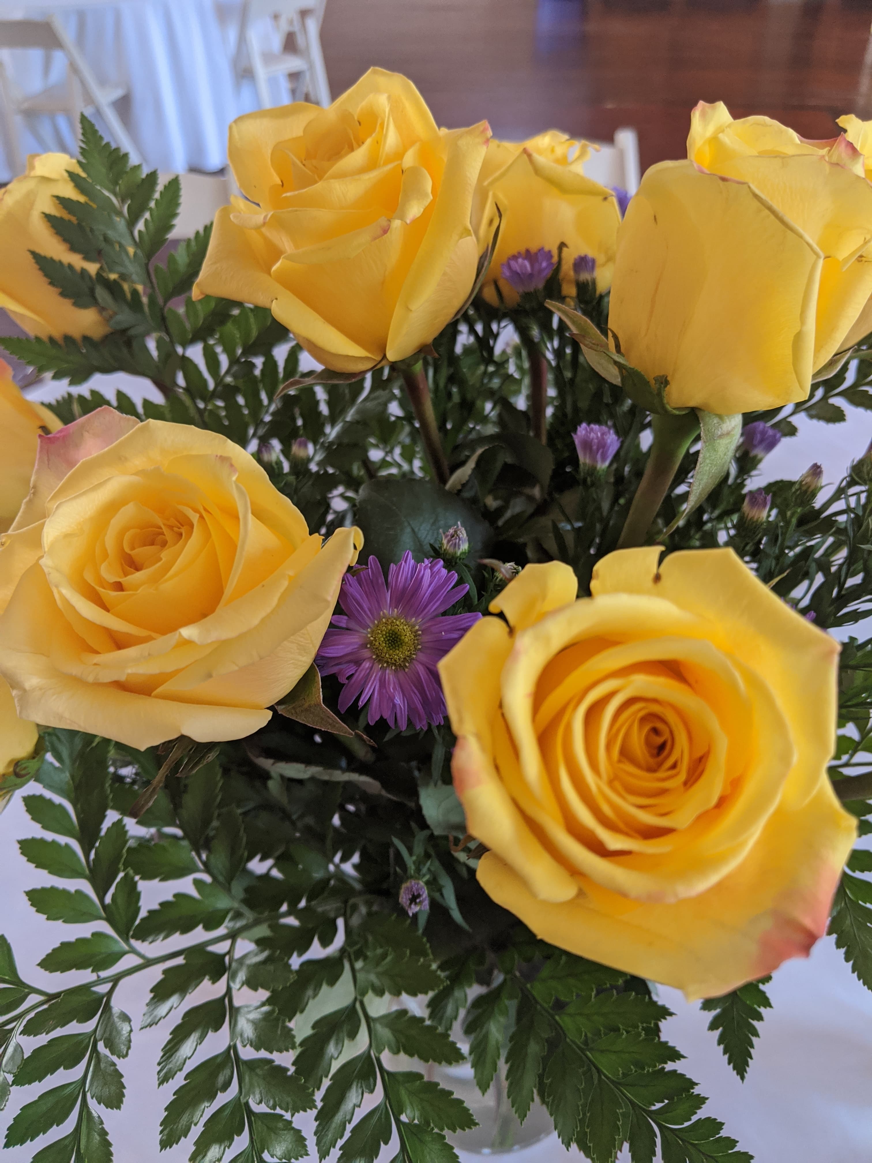 A close-up of a floral arrangement featuring bright yellow roses and a purple flower surrounded by green leaves.