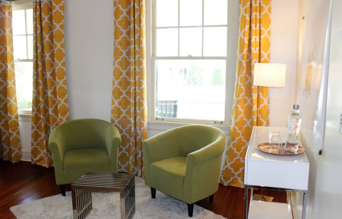 A cozy seating area with two green chairs, a striped side table, and bright yellow patterned curtains.