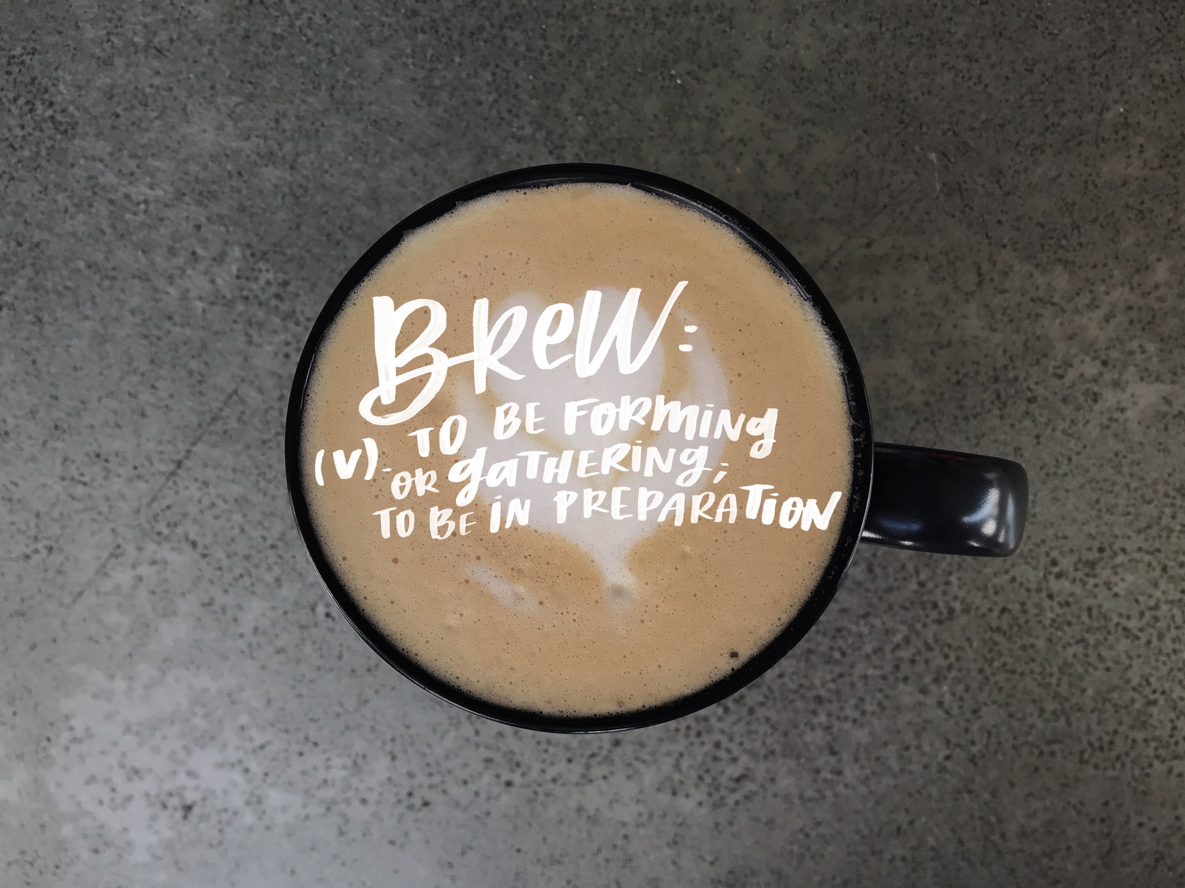 A close-up of a mug of coffee with the text "Brew: (v.) to be forming or gathering; to be in preparation" written on top.