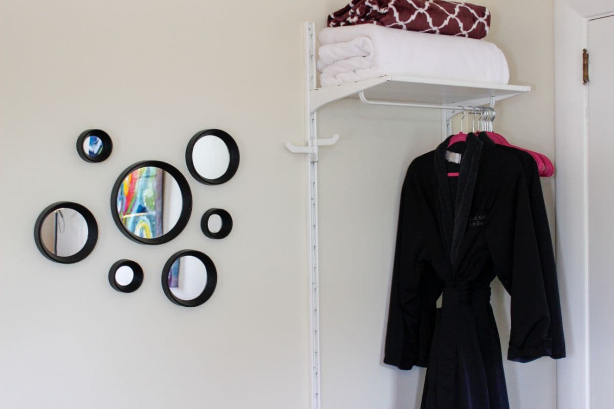 A black robe hangs next to a set of decorative mirrors and neatly stacked towels on a white shelf.