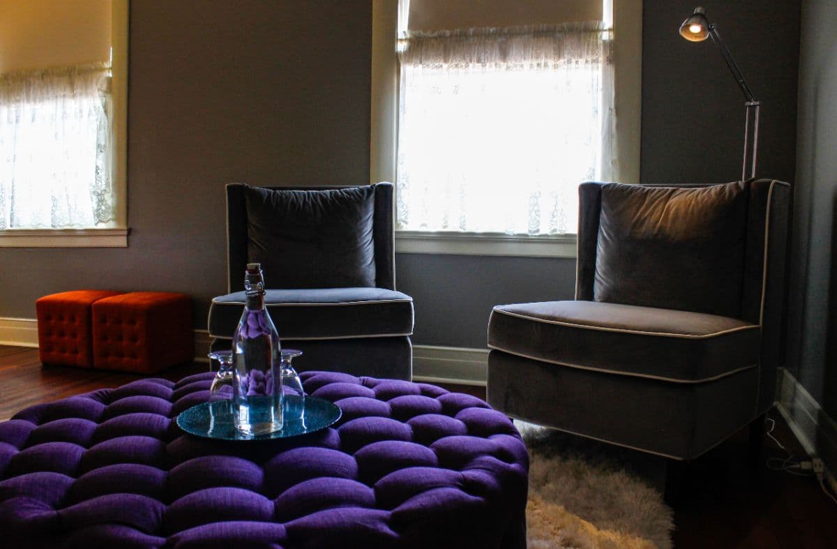 A cozy seating area featuring two gray chairs, a purple tufted ottoman, and a small orange cube, illuminated by natural light.