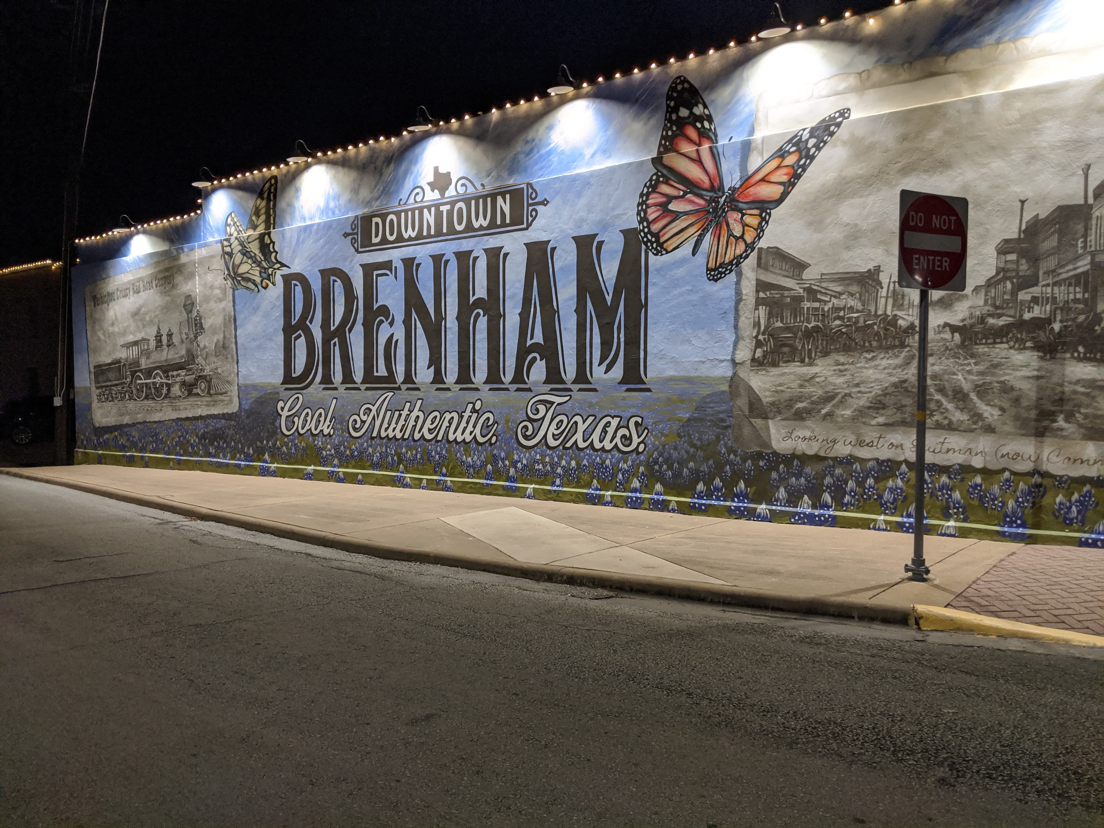 A vibrant mural featuring "Downtown Brenham, Cool Authentic Texas," with butterflies and historical images, illuminated at night.