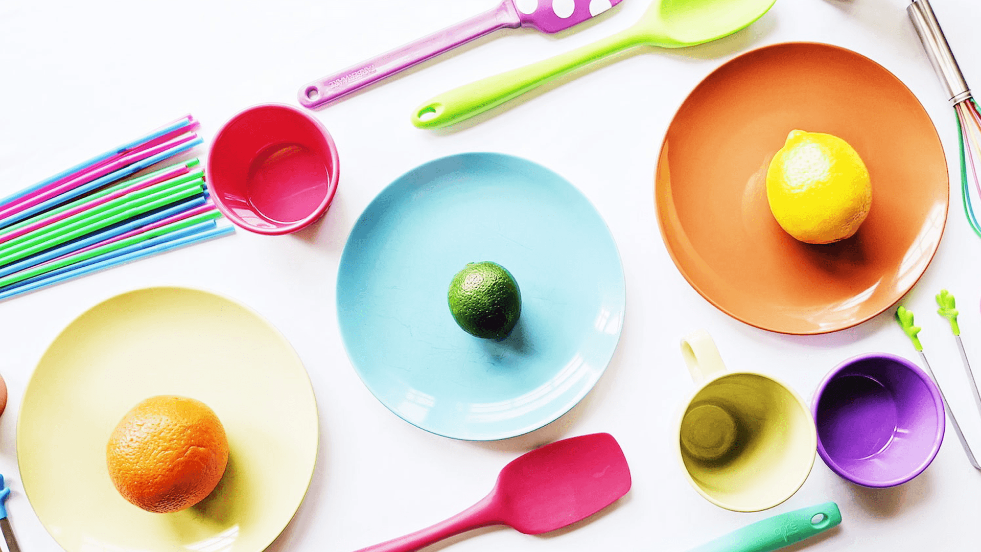 A colorful arrangement of plates, cups, utensils, and assorted fruits on a white surface.