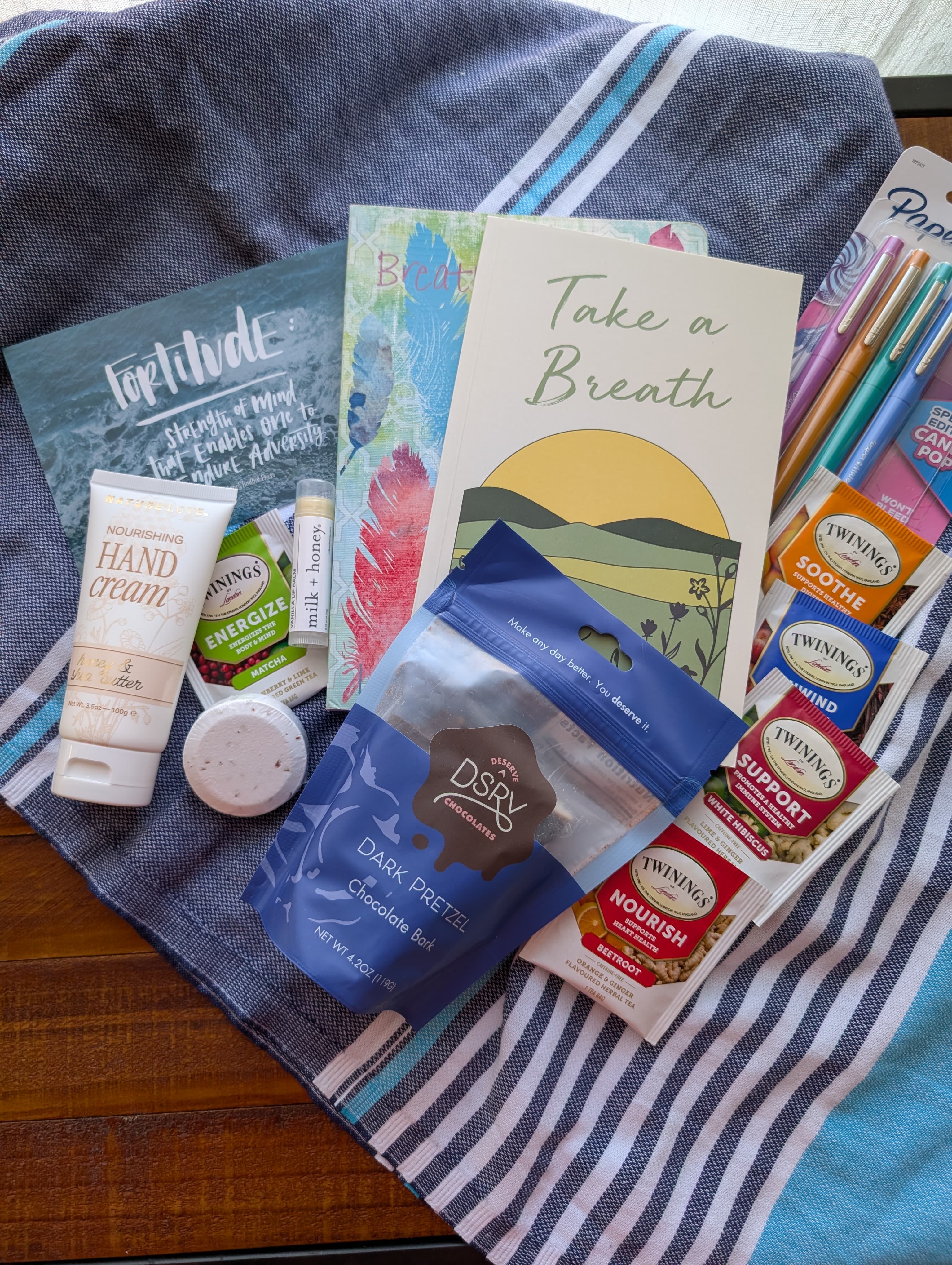 A cozy collection of self-care items including hand cream, herbal teas, a journal, and dark pretzels on a striped cloth.