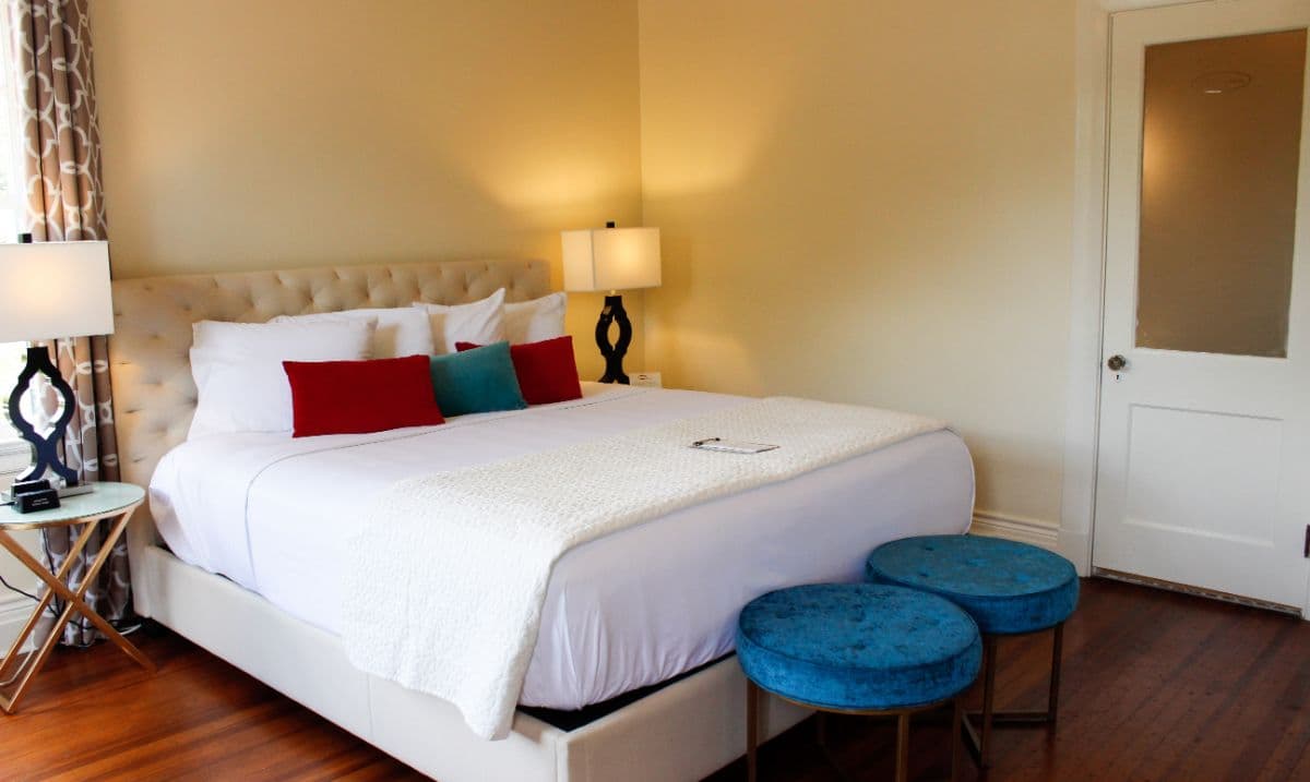 A cozy bedroom featuring a large bed with colorful pillows, two blue stools, and bedside lamps.