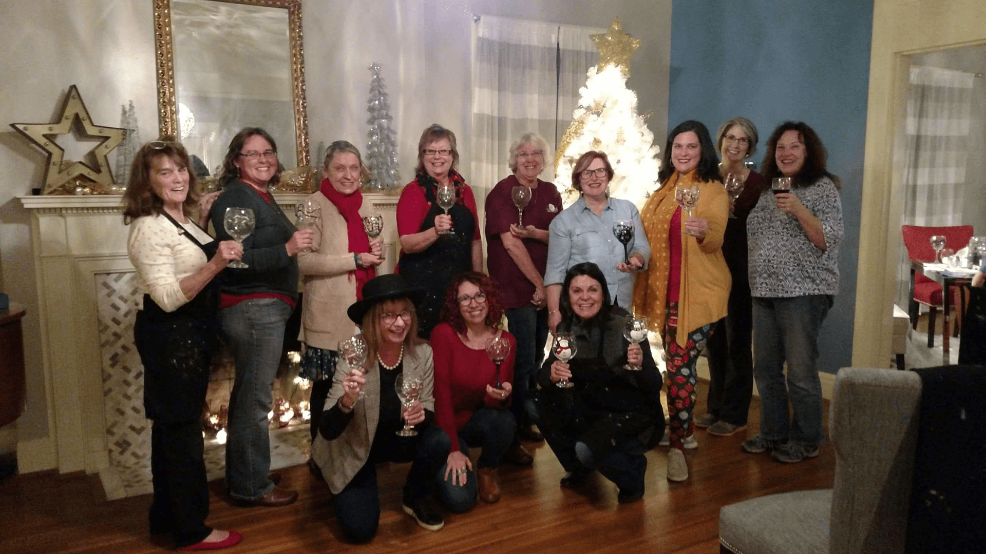 A group of twelve women cheerfully pose together, holding glasses, in front of a decorated Christmas tree.