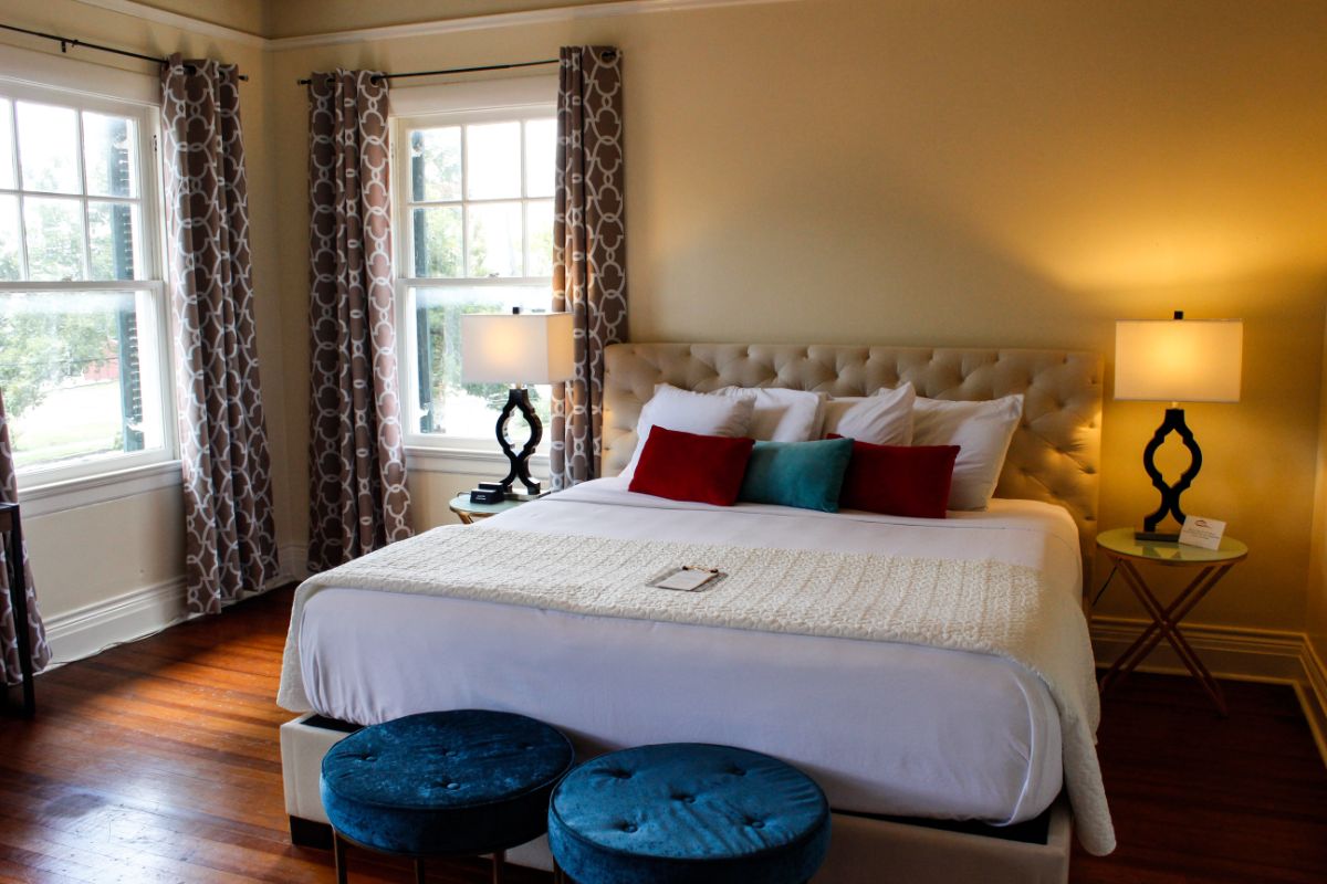 A cozy bedroom featuring a lavish bed with colorful pillows, two blue stools, and large windows with patterned curtains.