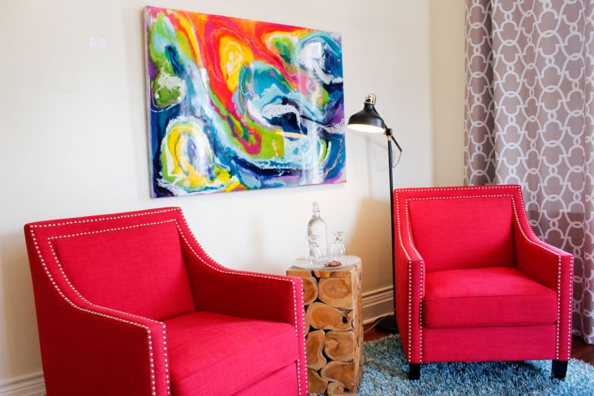 Two red armchairs face a colorful abstract painting, with a wooden side table and a lamp nearby.