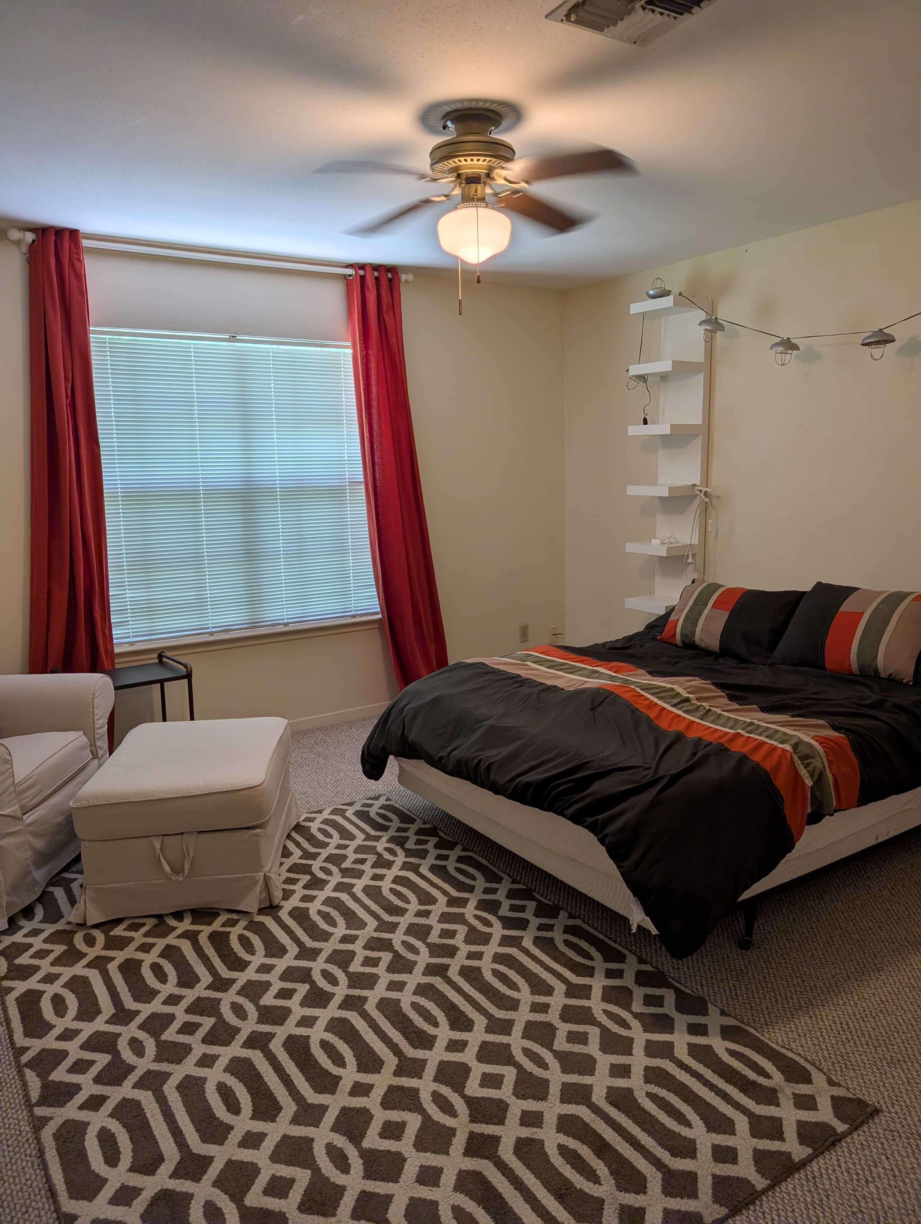 A cozy bedroom featuring a bed with dark bedding, a window with red curtains, a chair, and a patterned rug.