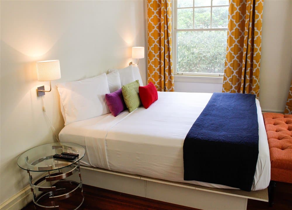 A cozy bedroom featuring a double bed with colorful pillows, a glass side table, and bright patterned curtains.